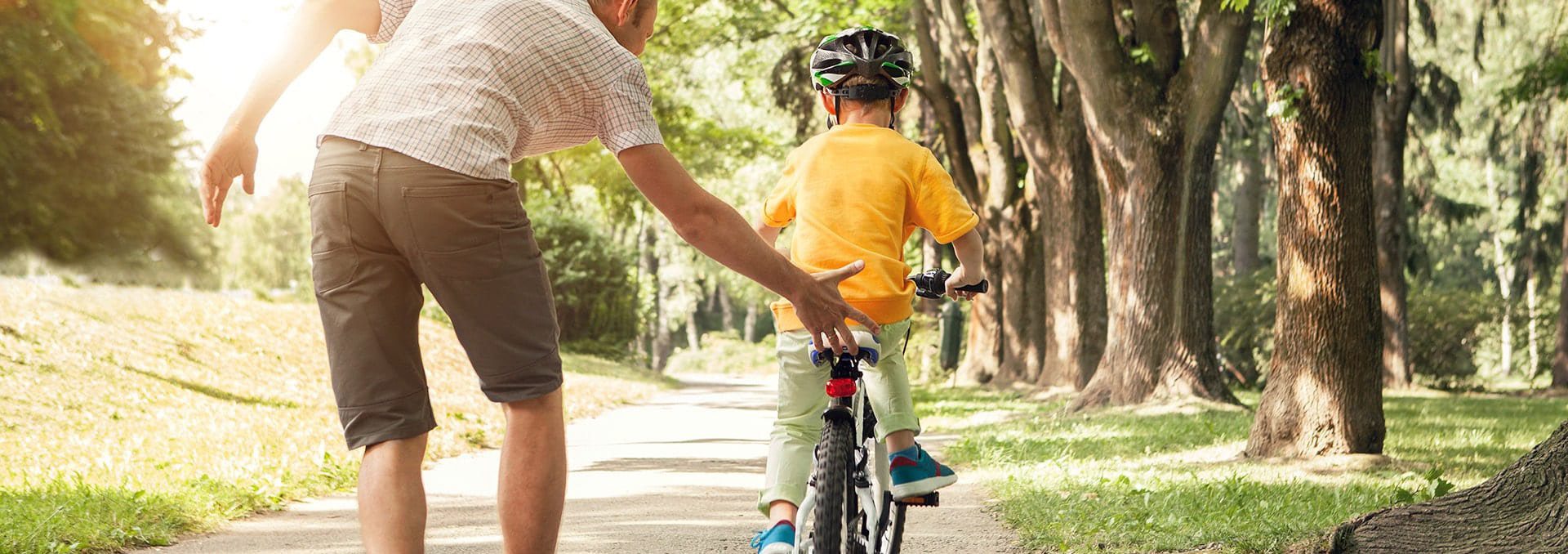 Father on tree-lined sidewalk teaching child to ride a bike