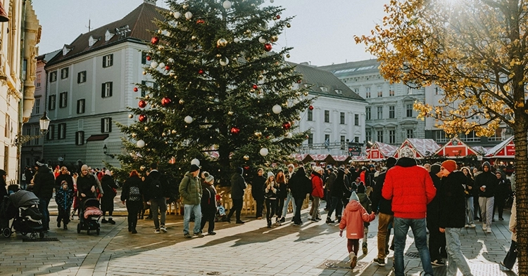People gathered in a town square around a large decorated Christmas tree near holiday market booths.